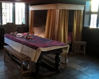 A four poster bed in the background and a table with a bench in the foreground of a historical room on Oak House Museum