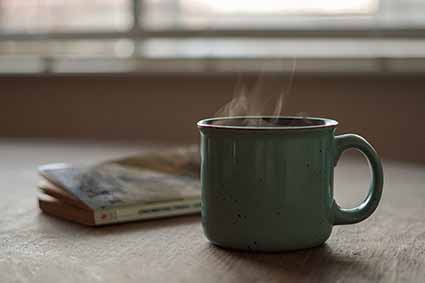A green mug next to a book on a table in front of a window, with steam coming from the liquid in the mug.