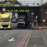 Two lorries passing under a low metal bridge.