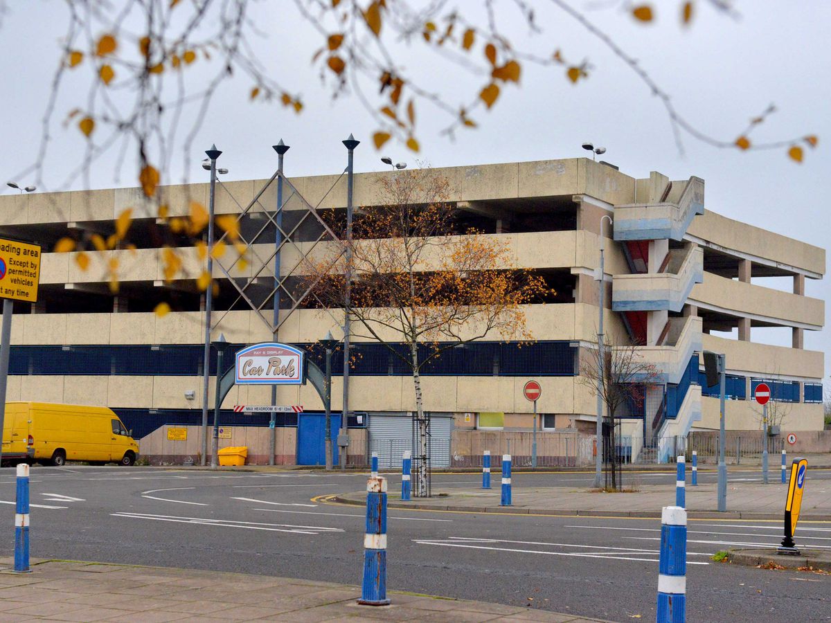 A multi-storey car park with a yellow van parked in front and tree branches hanging down in the foreground.