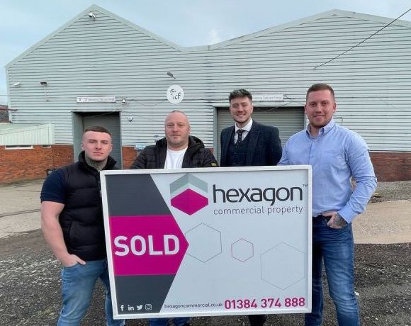 Four men standing in front of an industrial warehouse and holding up a sign saying 'sold'.