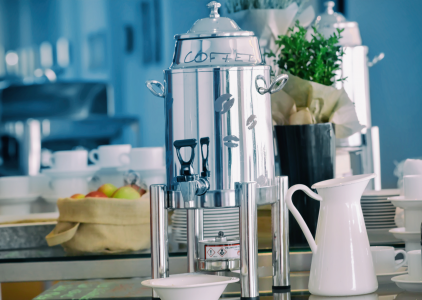 A polished silver tea urn next to a white jug and some piles of cups and saucers on a table with a basket of fruit in the background