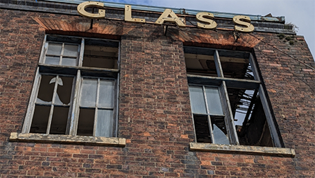 Close up of a derelict red brick building with broken windows and a sign saying Glass