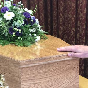 Close up of a hand on a wooden coffin with white and purple flowers on top