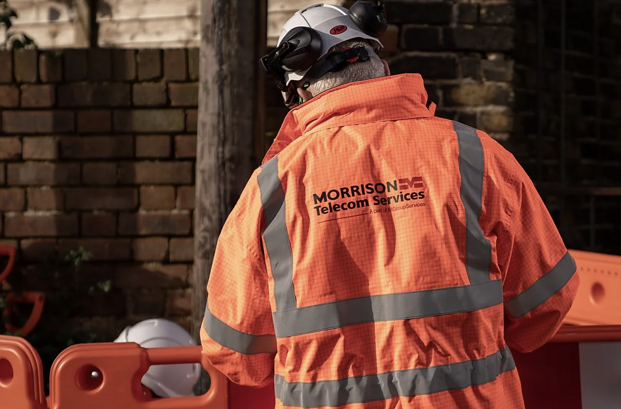 The back of a workman wearing a high vis orange workwear jacket and hard hat, standing in front of a wall