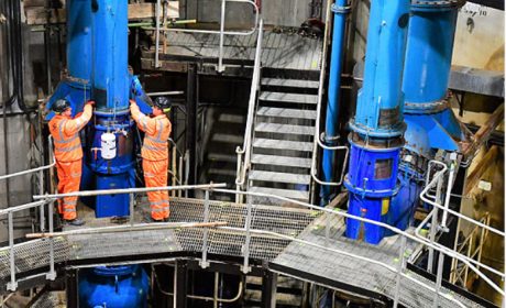 Two workers wearing orange PPE and hard hats working on a platform in a factory setting 