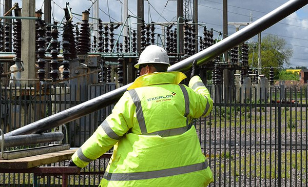 Workman wearing a hard hat and fluorescent yellow coat in front of some electricity pylons