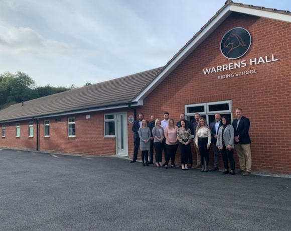 A group of thirteen office workers standing outside a red brick building with signage saying 'Warrens Hall'