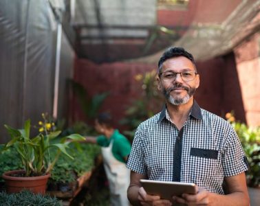 A man standing in a greenhouse, holding an iPad and looking towards the camera.