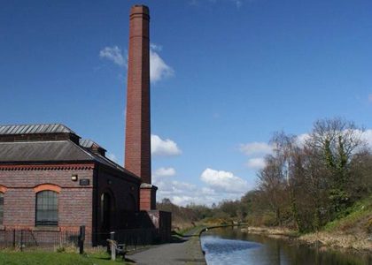 A Black Country local heritage site - a tall brick chimney and building next to the tow path of a canal with trees on the other side of the canal and blue sky in the background.