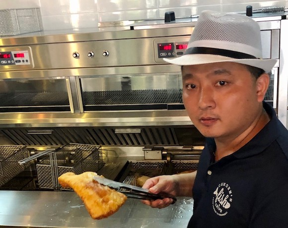 A man in a food preparation safety hat standing next to a commercial fryer holding a piece of battered, fried fish in a pair of tongs.