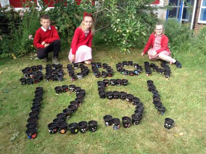 Three school children in red jumpers sitting on the grass next to a display of pots spelling out "Seddon 125!"