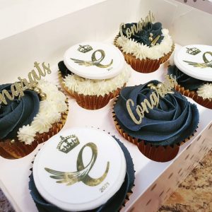 A box of six cupcakes topped with blue and white icing and gold decorations saying "Congrats"