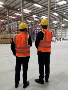 Two people wearing suits with orange high-vis vests over the top and yellow hard hats standing in a large warehouse space with their backs to the camera.