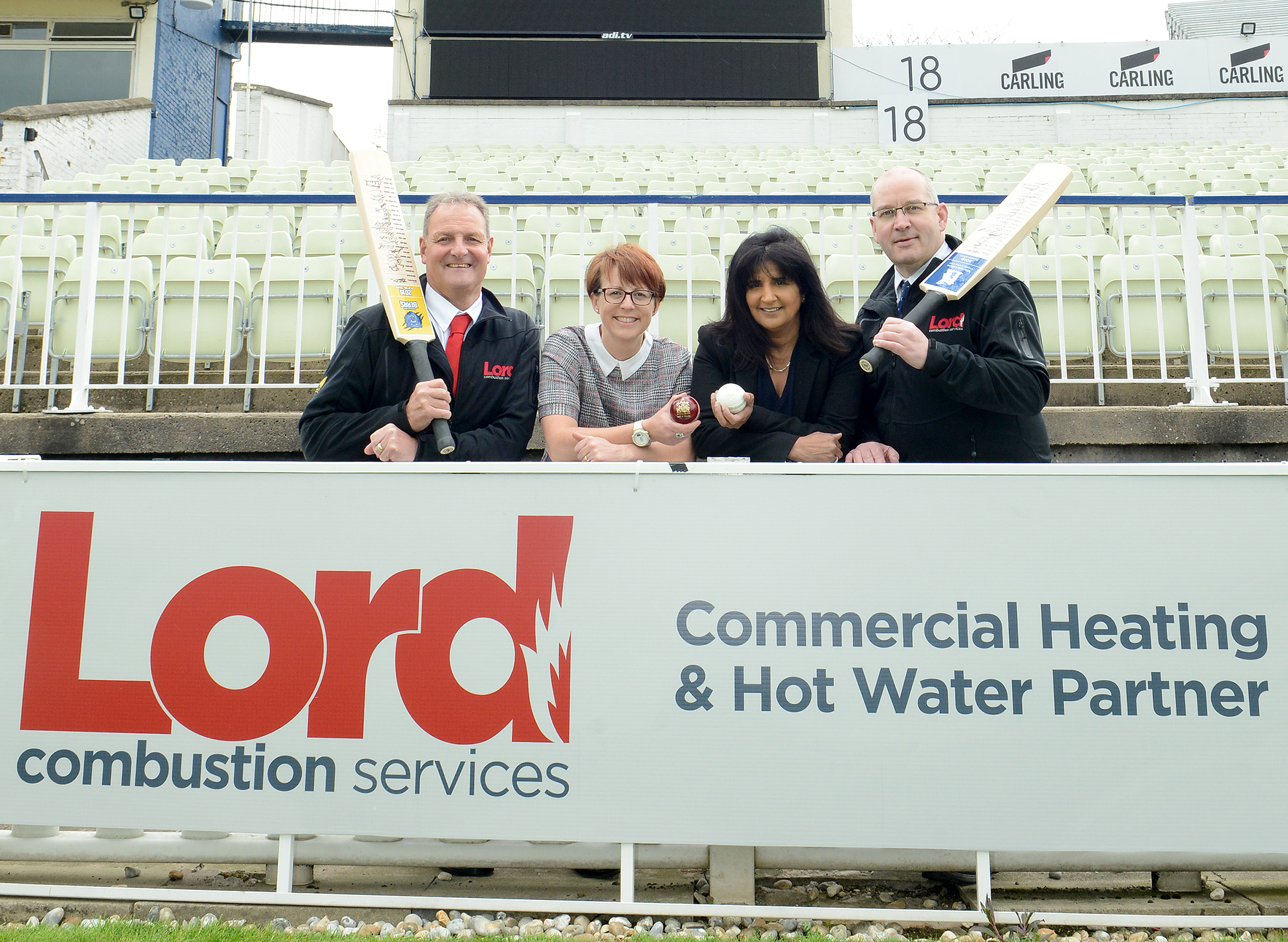 Four people standing behind a banner with the Lord Combustion Services logo on it, two of them holding cricket bats