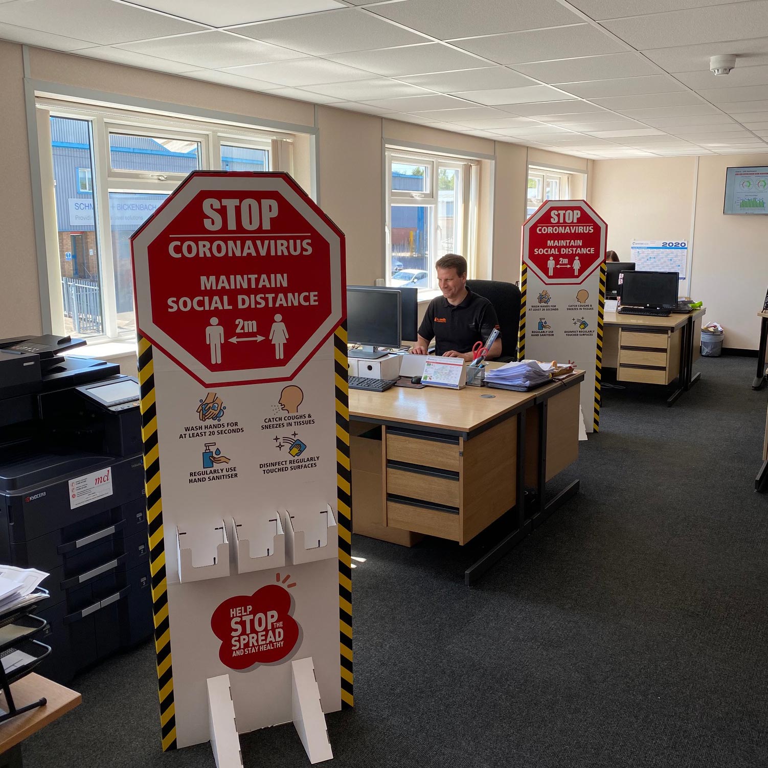 An office with a person sitting at a desk in the middle ground and a Stop sign in the foreground saying "Stop, Coronavirus - Maintain Social Distance"