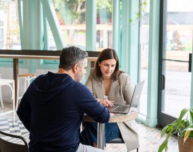 A white man in a dark blue jumper and a white woman in a grey jacket are sitting opposite each other at a table in a modern building, looking at an open laptop.