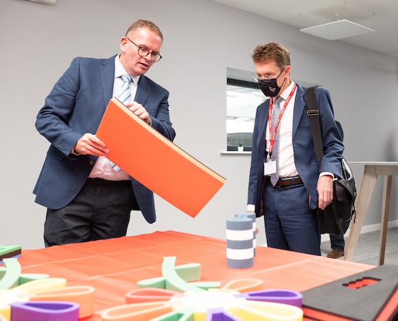 Two men in dark grey suits. One is showing the other a sheet of orange foam.