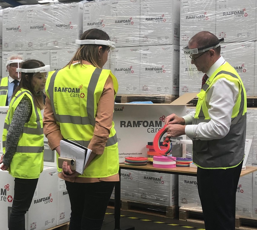 Three people wearing yellow high-vis vests and plastic visor face masks standing infront of a large pile of boxes.