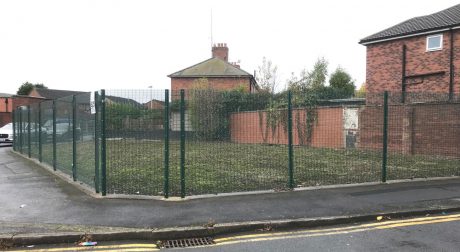 An area of grass on a street corner with a metal fence around it and red brick houses in the background