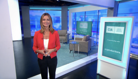 Screenshot of a YouTube video showing a female newsreader wearing a red jacket standing in front of some screens