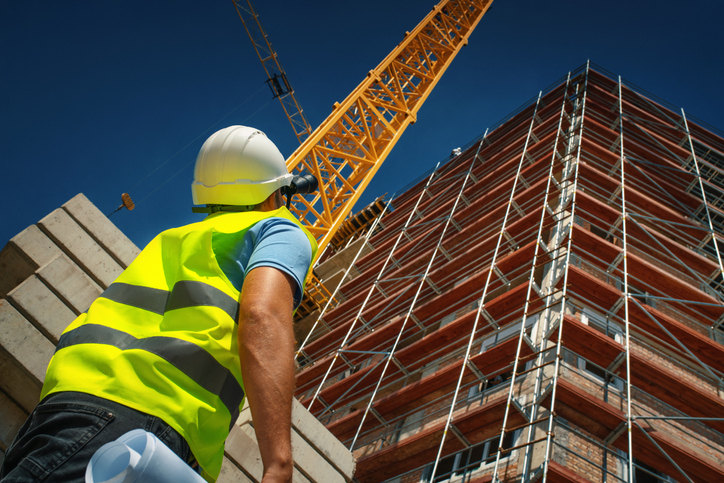 A person in a high vis jacket and hard hat looking up at a tall building covered in scaffolding and crane next to it