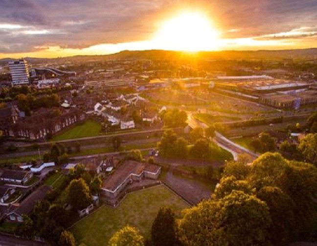 The sun setting in the background, casting a bright yellow light over houses and fields with trees in the foreground.