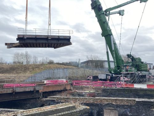 A green crane lifting up part of a metal bridge.