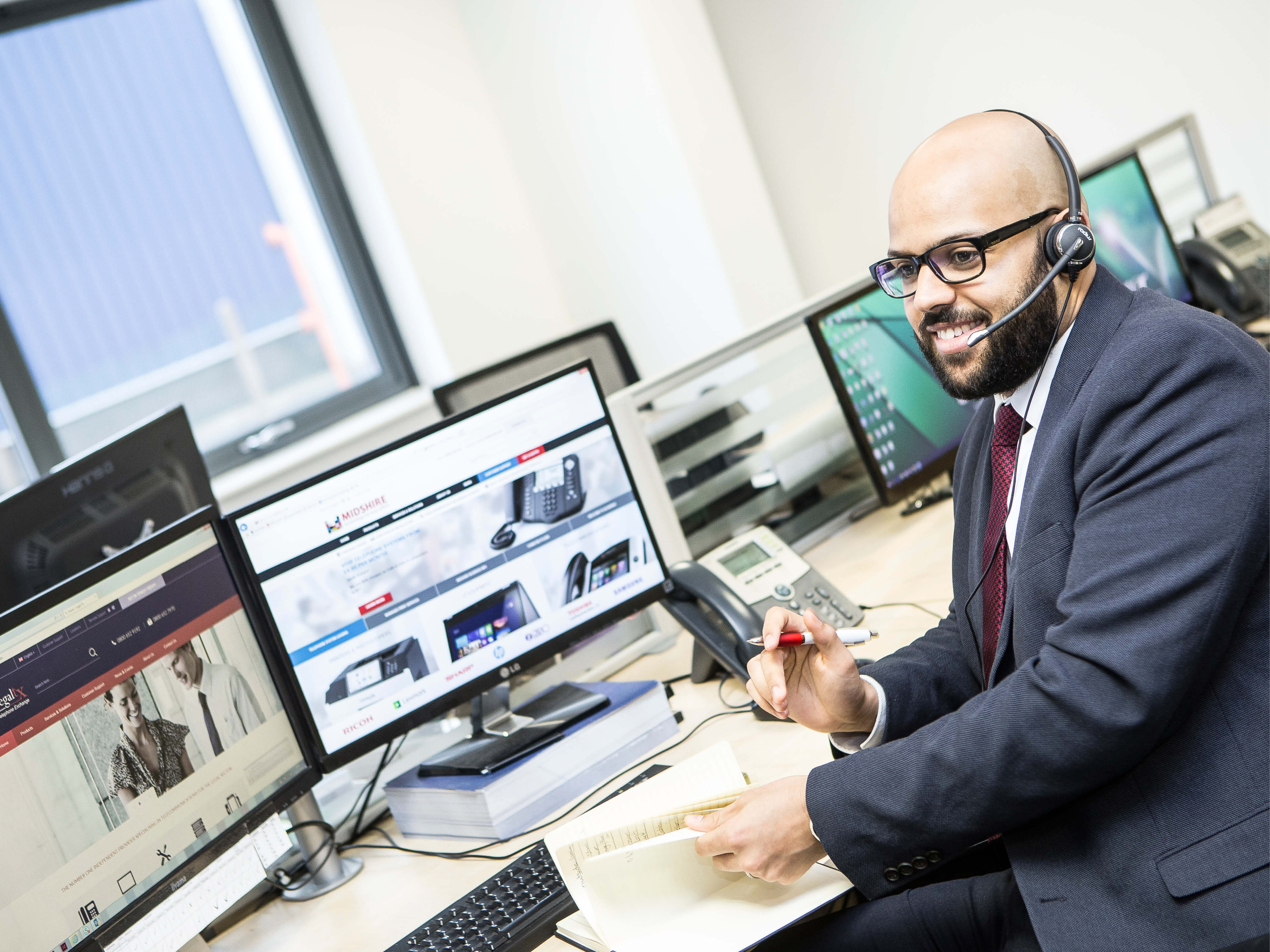 Man with a pen and a book at a desk of computers, smiling and speaking into a headset