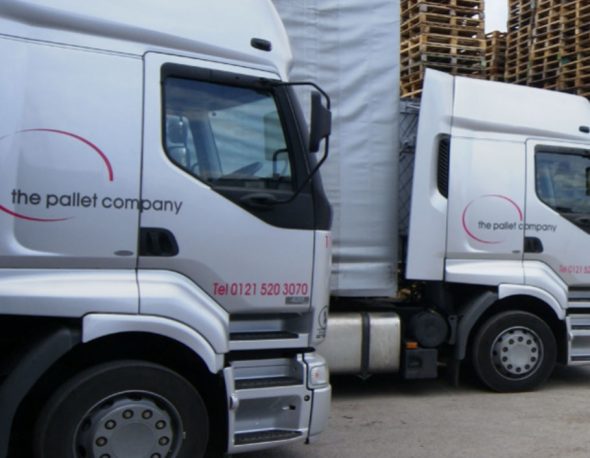 The cabs of two Pallet Company lorries parked infant of a tall stack of wooden pallets.