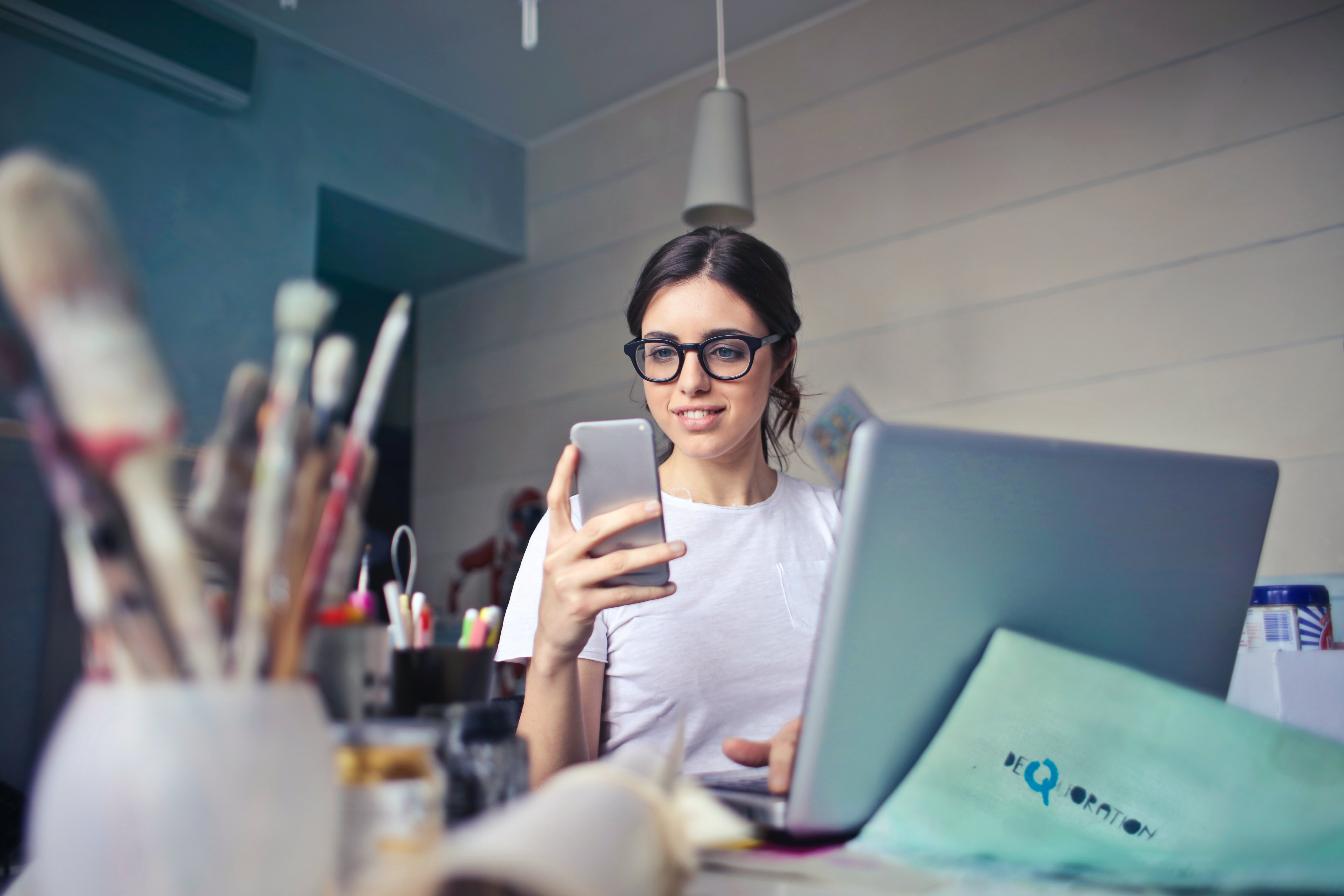 A young woman wearing glasses sitting at a laptop holding a mobile phone in front of her, with a pot of paint brushes out of focus in the corner of the foreground