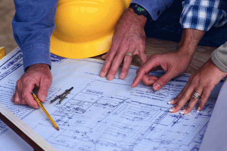 Close up of architect's plan with a compass and yellow hard hat on it and four hands pointing at the drawing.