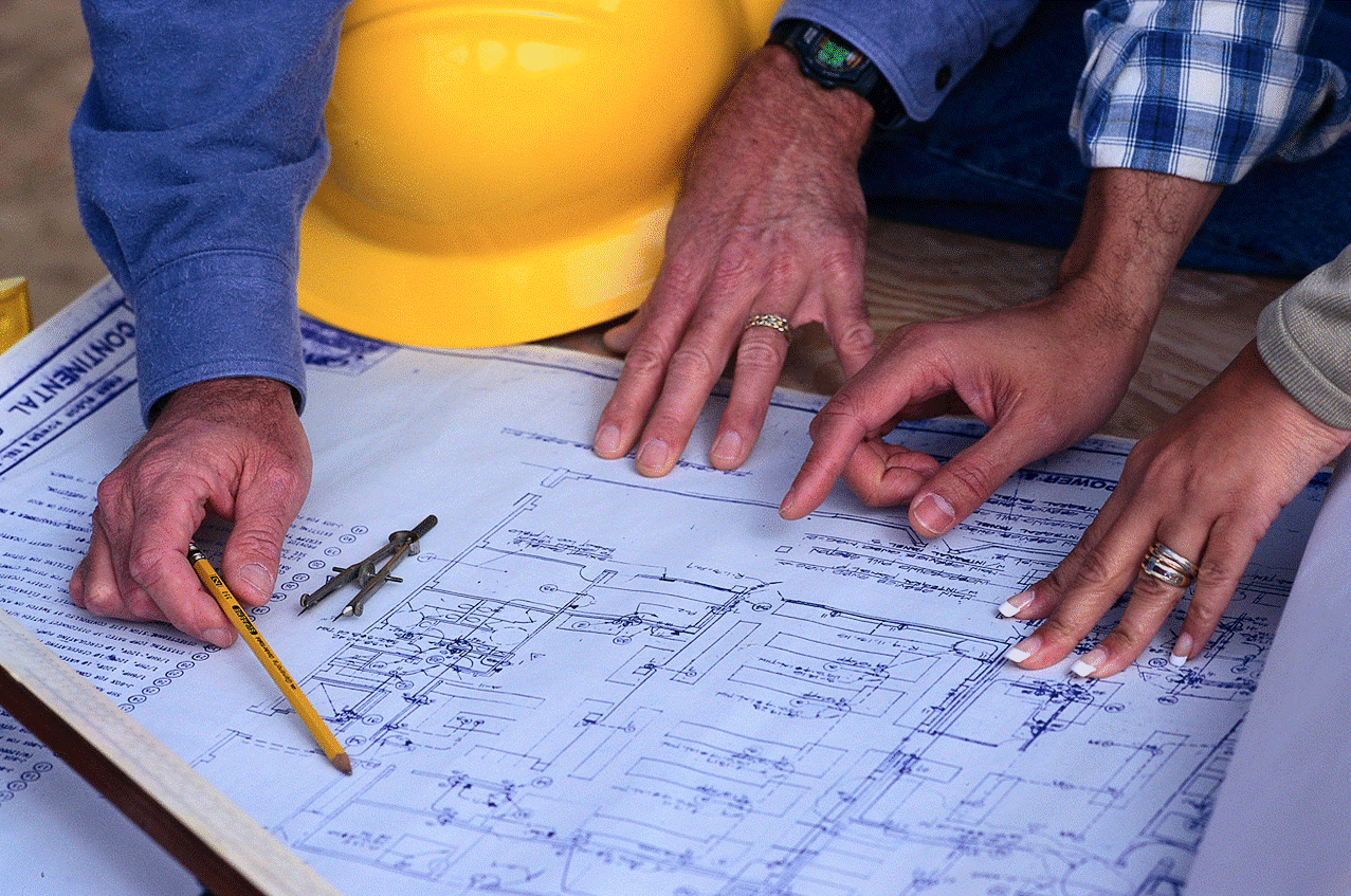 Close up of architect's plan with a compass and yellow hard hat on it and four hands pointing at the drawing.