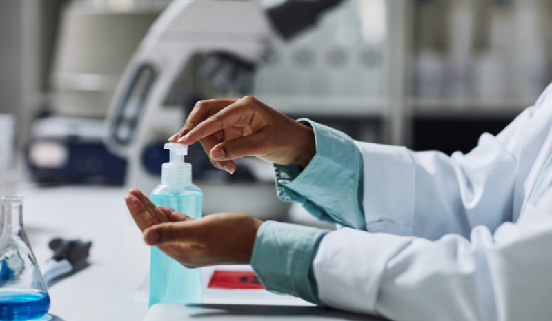 A close-up of a woman's hands - slim, brown skin, wearing a lab coat or overalls - applying hand sanitiser at a workstation