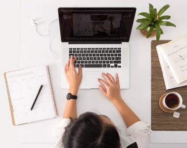 A photograph from above of a person sitting at a white desk with a notebook and pen, open laptop, small plant, two books and a cup of tea.