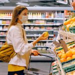 A woman wearing a mask holding a basket in one hand and an orange in the other, standing in front of the fruit and veg in a supermarket