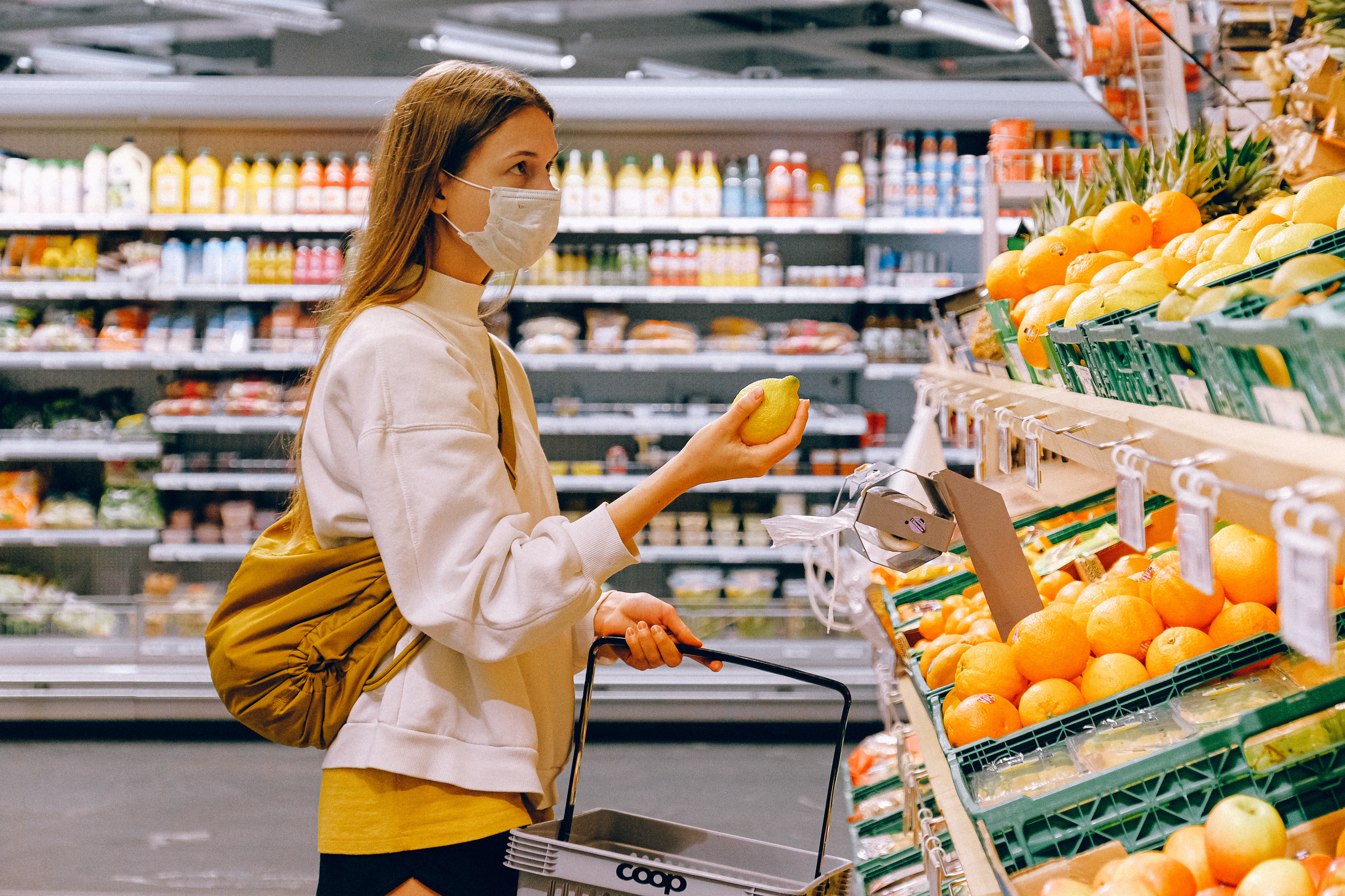 A woman wearing a mask holding a basket in one hand and an orange in the other, standing in front of the fruit and veg in a supermarket